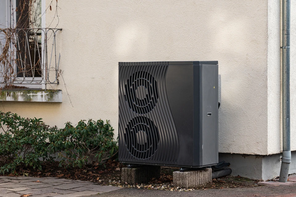 Photographie réaliste d'une pompe à chaleur air-eau installée à l'extérieur d'une maison résidentielle, montrant clairement l'unité compacte avec ses ventilateurs et son design moderne, en cours de fonctionnement ou fraîchement installée, dans un environnement résidentiel français typique avec maison individuelle. L'image doit mettre l'accent sur l'installation extérieure et le système de captage de l'air.