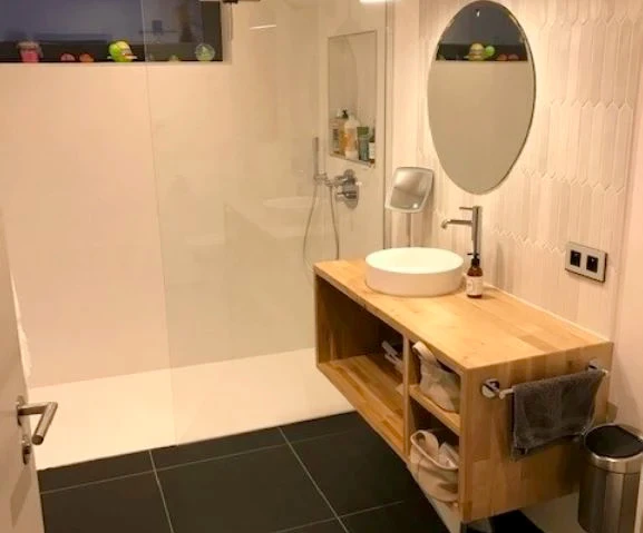 Deux plombiers en salopette bleue et casquette installent un lavabo vasque rond blanc sur plan de travail bois dans une salle de bain moderne